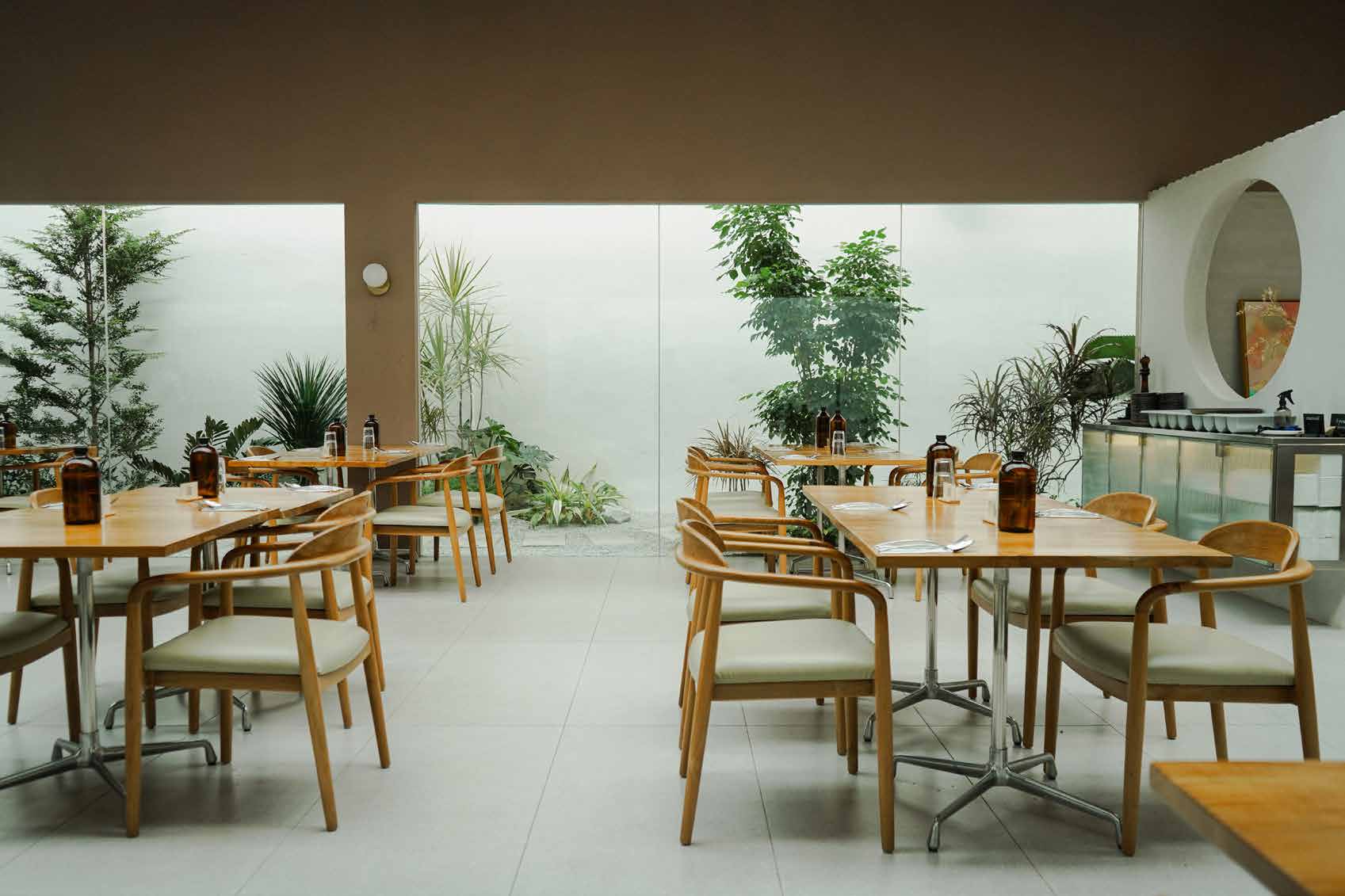 The Cafe Area: light wood chairs on white tiles, a round window and greenery behind glass
