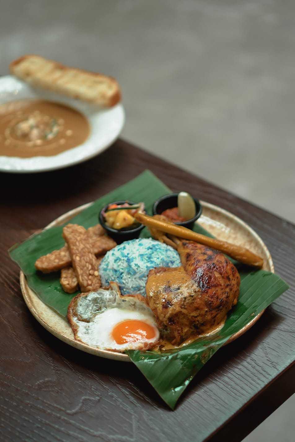 Nyonya-spiced roasted chicken leg on a banana leaf with blue pea rice, a fried egg and sambal
